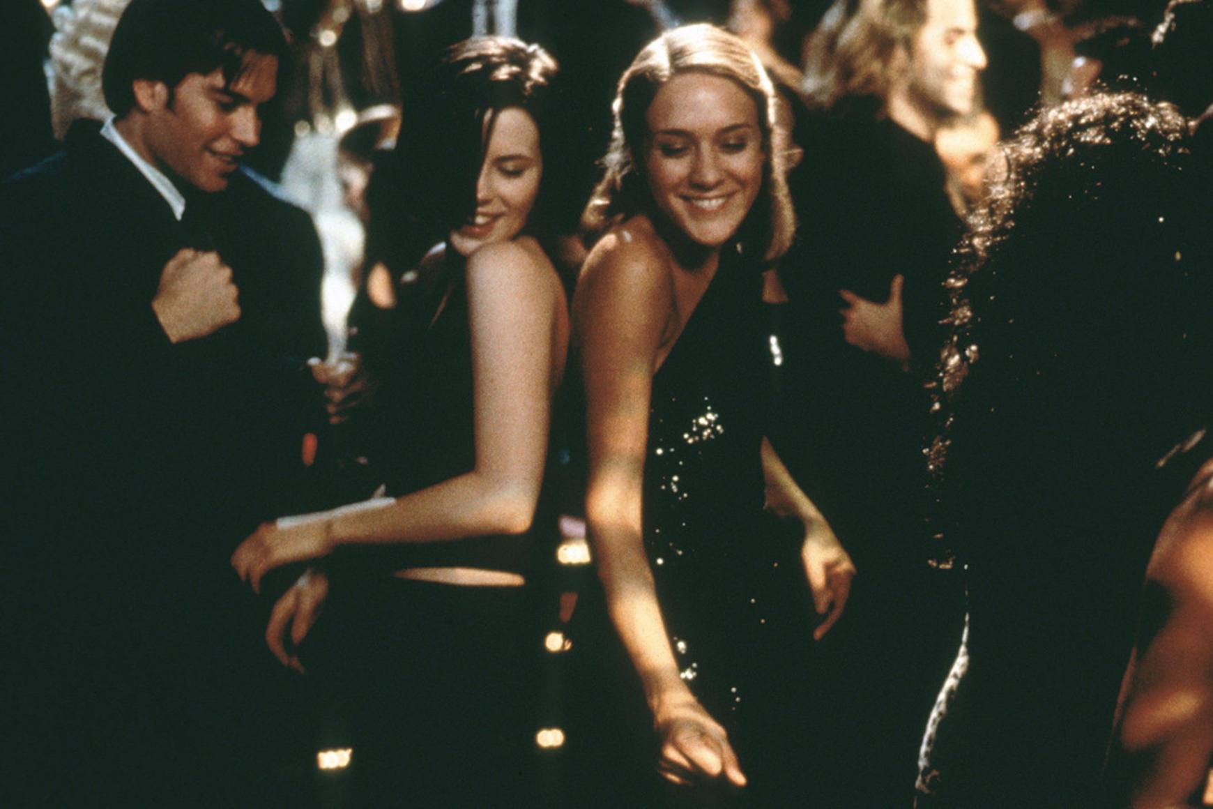 Kate Beckinsale and Chloë Sevigny smile and dance in black dresses in a crowded room. 